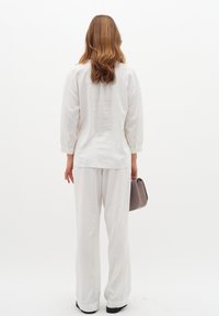 Blusa bianca in lino con maniche ampie, abbinata a pantaloni bianchi larghi. Gli accessori includono una piccola borsa grigia. Sfondo semplice.