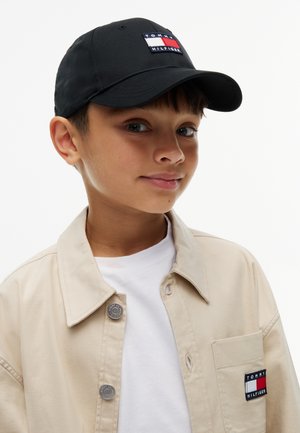 Niño con una chaqueta beige de Tommy Hilfiger sobre una camisa blanca y una gorra de béisbol negra de Tommy Hilfiger, mirando ligeramente hacia un lado.