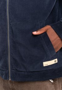 Navy corduroy jacket with a zip closure, side pockets, and a label featuring "EST. 1992" in cream. Textured fabric shows vertical lines.