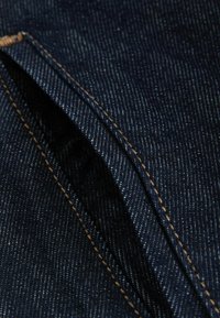 Tejido de mezclilla azul oscuro con una fina textura vertical. Presenta costuras amarillas a lo largo de las costuras y una abertura para un bolsillo lateral.