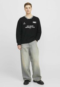 Sort strikket sweater med v-hals og hvide striber, prydet med grafiske prints og tekst. Båret med løse, lysegrå jeans og sorte sko.
