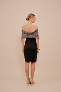 Sieviete ar matiem uz kakla, tērpusies melnā off-shoulder kleitā ar sudraba spīdīgu topu un bēšiem augstiem zābakiem, stāvot ar muguru vērsta uz priekšu.