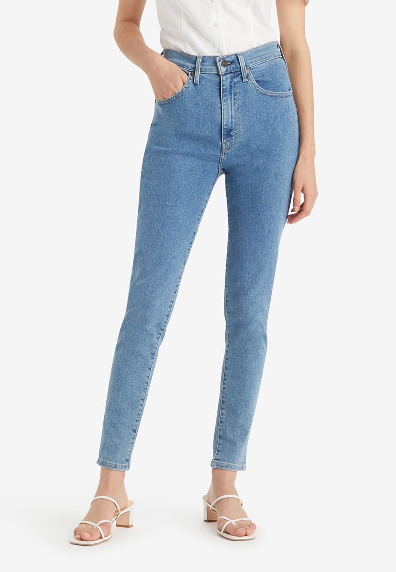 Femme portant un jean skinny taille haute bleu clair avec poches avant, associé à des sandales blanches à talons et à brides ainsi qu'une chemise blanche à manches courtes.