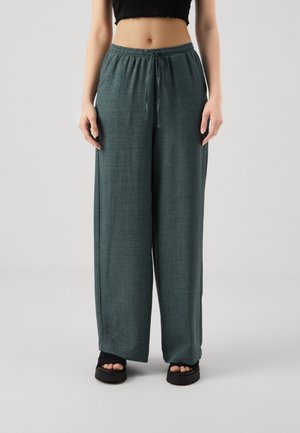 Personne portant un pantalon ample vert foncé à cordon et des sandales noires à bouts ouverts, debout devant un fond clair uni.