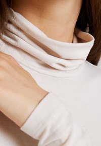 Helle beige Rollkragenoberteil aus einem weichen, dehnbaren Material mit umgeschlagenem Kragen und enganliegenden langen Ärmeln. Subtile Nahtdetails sind sichtbar.