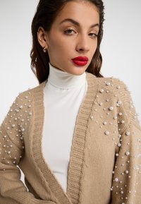 Beige pletený kardigan s perlovými akcenty na ramene, s otevřeným předním dílem a žebrovanou texturou. Nosený přes bílý rolák.