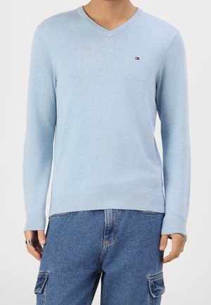 Man draagt een lichtblauwe V-hals trui met een klein logo op de borst en blauwe cargobroek, staand met ontspannen handen langs het lichaam.