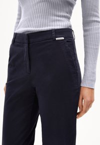 Navyblauwe katoenen broek met een gladde voorkant, zijzakken en een klein merkembleem nabij de tailleband. Geribbelde grijze trui zichtbaar aan de bovenkant.