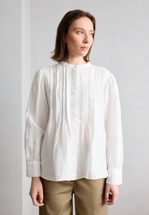 Γυναίκα με καστανά μαλλιά μήκους μέχρι τους ώμους, που φοράει μια λευκή μπλούζα με μακριά μανίκια και πιέτες, και μπεζ παντελόνι, κοιτάζει προς τα δεξιά της.