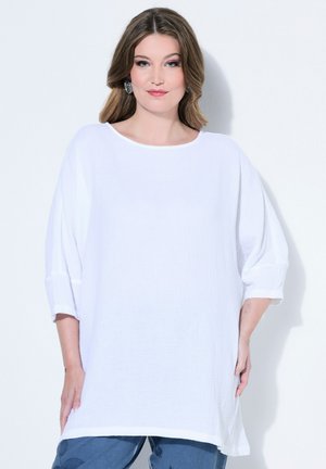 Vaaleanruskeat hiukset omaava nainen pukeutuneena ylisuureen valkoiseen, teksturoituun paitaan, jossa on kolme neljäsosahihat ja sinisiin, kuviollisiin housuihin, seisoo valkoista taustaa vasten.