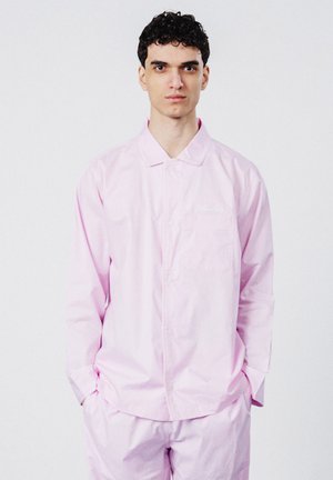 Junger Mann mit lockigem dunklem Haar trägt ein hellrosa Hemd und Hose, steht vor einem schlichten weißen Hintergrund, die Hände in den Taschen.