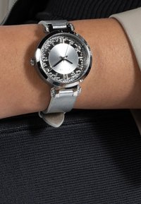 Sølvmetallur med rund urskive, funklende detaljer og en lys grå læderrem. Har en glat overflade og minimalistisk design.