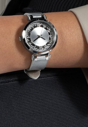 Orologio in metallo argentato con un quadrante rotondo, dettagli brillanti e un cinturino in pelle grigio chiaro. Presenta una finitura liscia e un design minimalista.