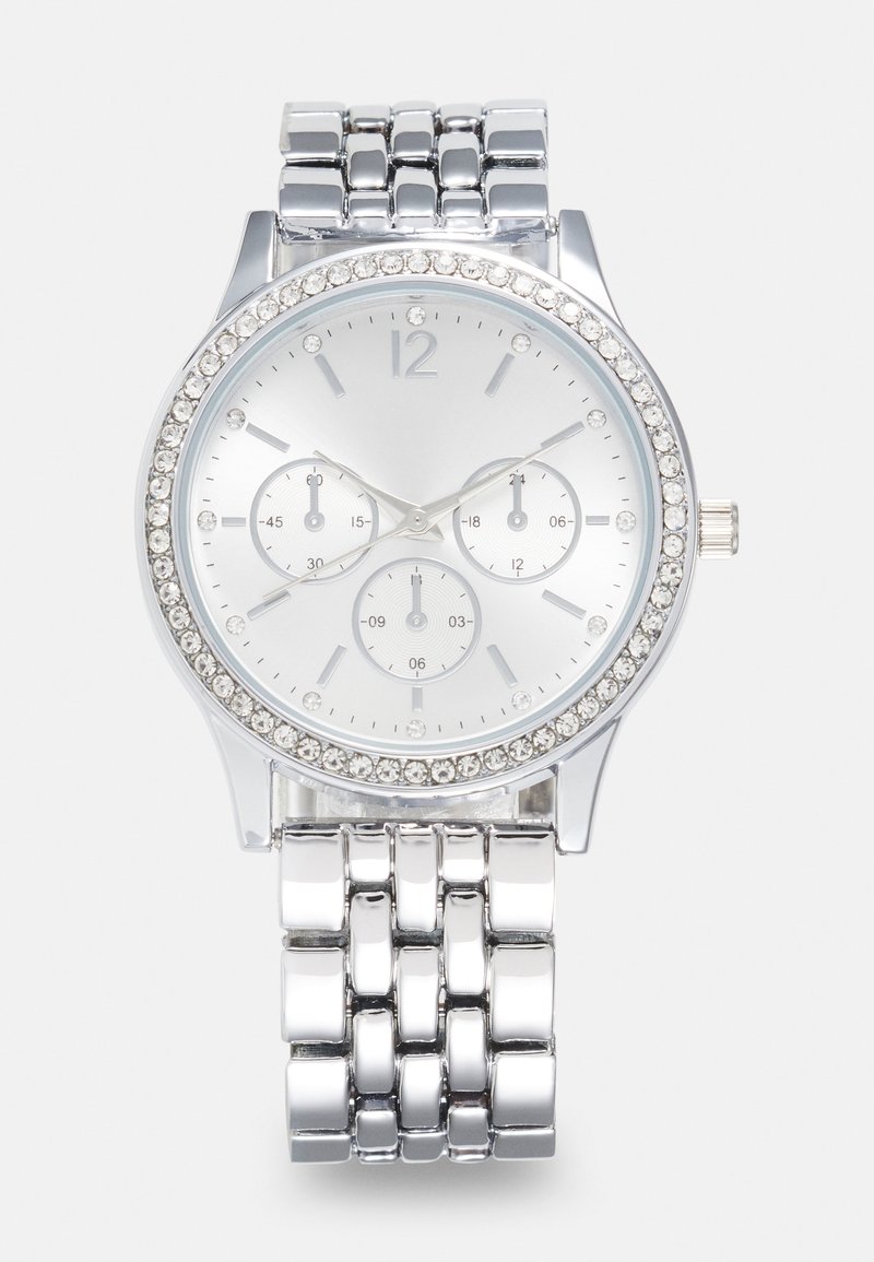 Montre analogique en argent avec un bracelet en métal brillant, un cadran rond et des accents en cristal autour de la lunette. Présente trois sous-cadrans sur fond blanc.