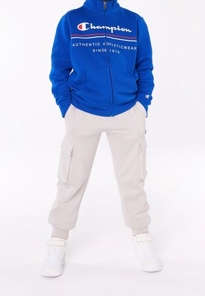 Blauer Reißverschluss-Pullover mit weißem Logo und rotem Akzent; beige Cargo-Sweatpants mit Seitentaschen; weiße Sportschuhe.