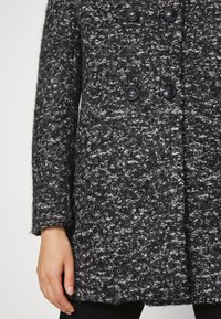 Ciemnoszary, teksturowany wełniany płaszcz z czarnymi guzikami, widoczny od połowy tułowia do nadgarstka, noszony przez osobę z jedną ręką swobodnie opuszczoną wzdłuż ciała.