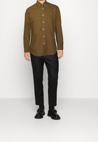 Camicia marrone con bottoni e colletto, abbinata a pantaloni neri e scarpe nere, caratterizzata da una vestibilità slim e un design minimalista.