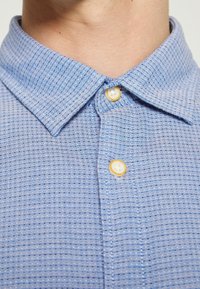 Gros plan d'une personne portant une chemise bleue à motifs avec les deux premiers boutons fermés.