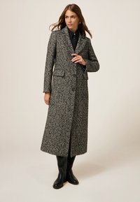 Cappotto a scacchi in bianco e nero, con maniche lunghe, colletto a rever e tasche frontali, abbinato a stivali neri al ginocchio.