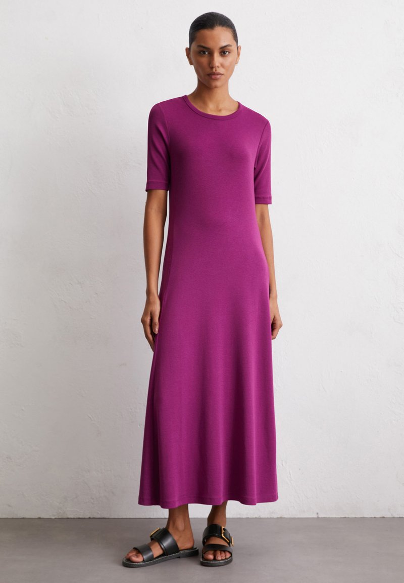 Lila knielanges Kleid mit kurzen Ärmeln, aus glattem Stoff. Einfaches Design mit Rundhalsausschnitt und fließender Silhouette, kombiniert mit schwarzen Sandalen.