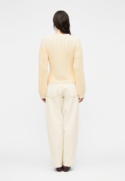 Γυναίκα με μακριά σκούρα μαλλιά που φοράει ένα κρεμ πλεκτό πουλόβερ με σχέδιο κόμπων και κρεμ παντελόνι ψηλόμεσο με φαρδιά μπατζάκια, στέκεται γυρισμένη πλάτη σε λευκό φόντο.