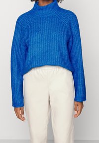 Blå stickad tröja med hög krage, ribbad textur och oversize passform, ihop med krämfärgade byxor.