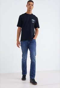 Camiseta de algodón negra de manga corta con un gráfico frontal, combinada con unos pantalones vaqueros azules y zapatillas negras con suelas marrones.