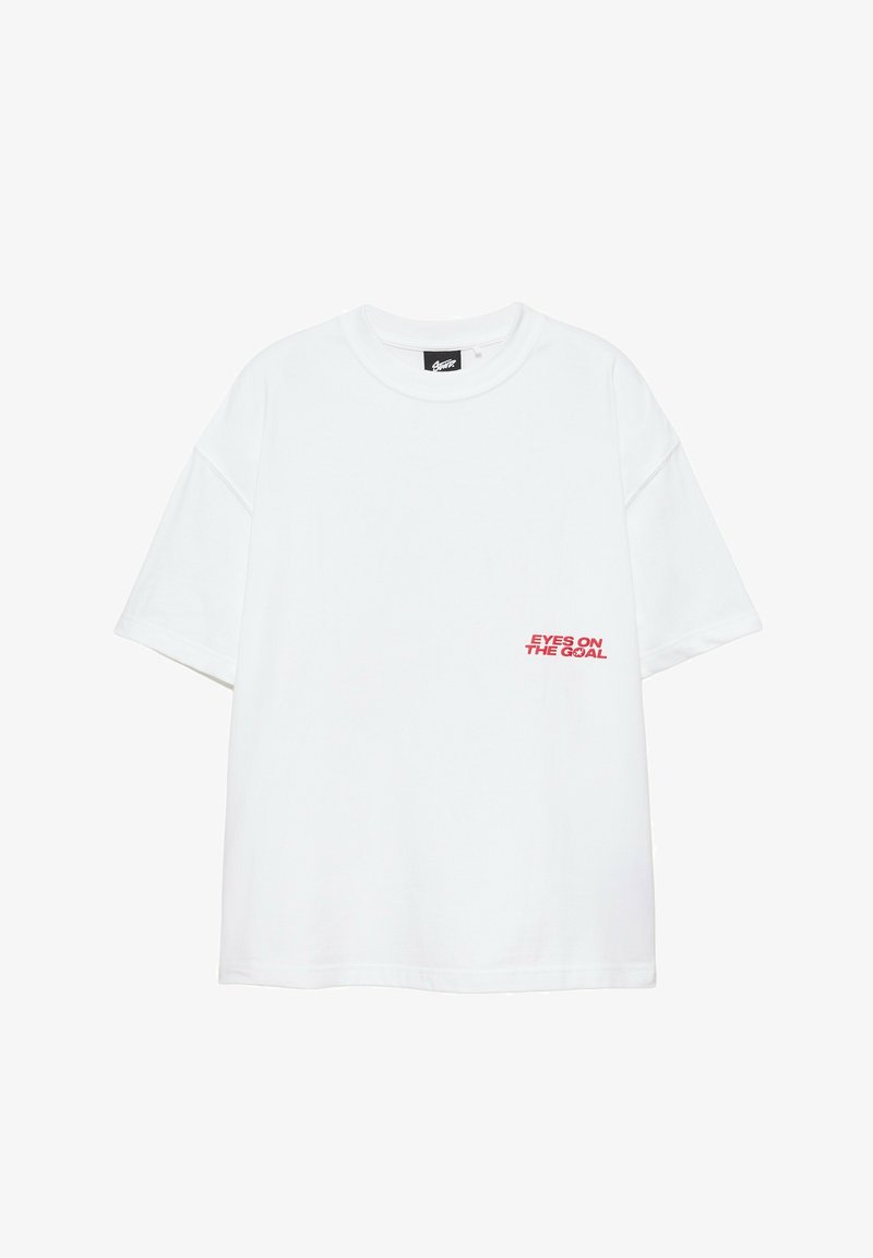 T-shirt blanc à manches courtes avec un texte rouge "EYES ON THE GOAL" en bas à droite sur le devant et une étiquette de marque noire à l'intérieur du col.