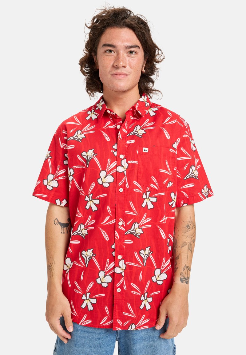 Camisa roja de manga corta con un patrón floral que presenta flores blancas y hojas verdes; diseño de botones y un bolsillo en el pecho.