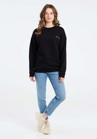 Felpa nera in misto cotone, con collo tondo, vestibilità relax e piccolo logo sul petto, abbinata a jeans blu e sneaker.