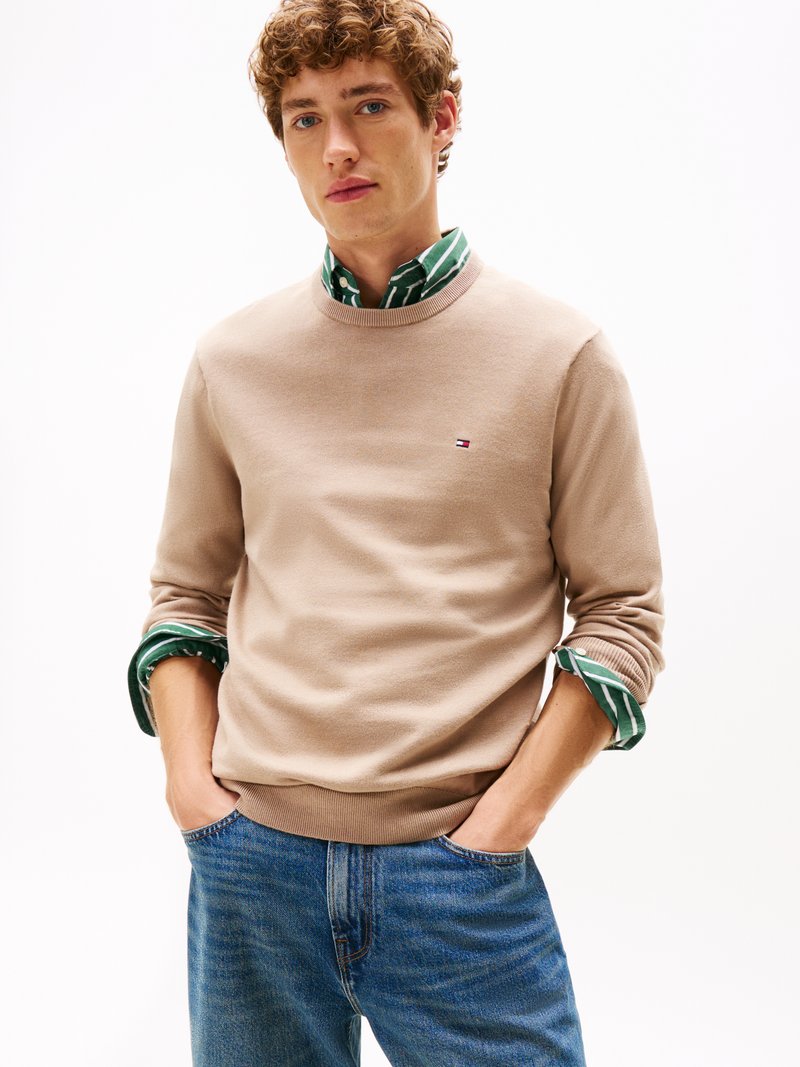 Beige pulover z okroglim izrezom in rebrastimi manšetami, položen čez zeleno in belo črtasto majico, v kombinaciji z modrimi jean hlačami.
