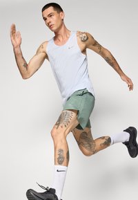 Hellblaues Tanktop und grüne Shorts, beide mit atmungsaktivem Design, schwarze Sportschuhe, weiße Socken und mehrere detaillierte Tattoos.