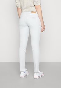 Světle modré skinny džíny s hladkým texturem, obsahující dvě zadní kapsy, surové lemování a hnědou koženou štítku v pase. Spárované s bílými teniskami.