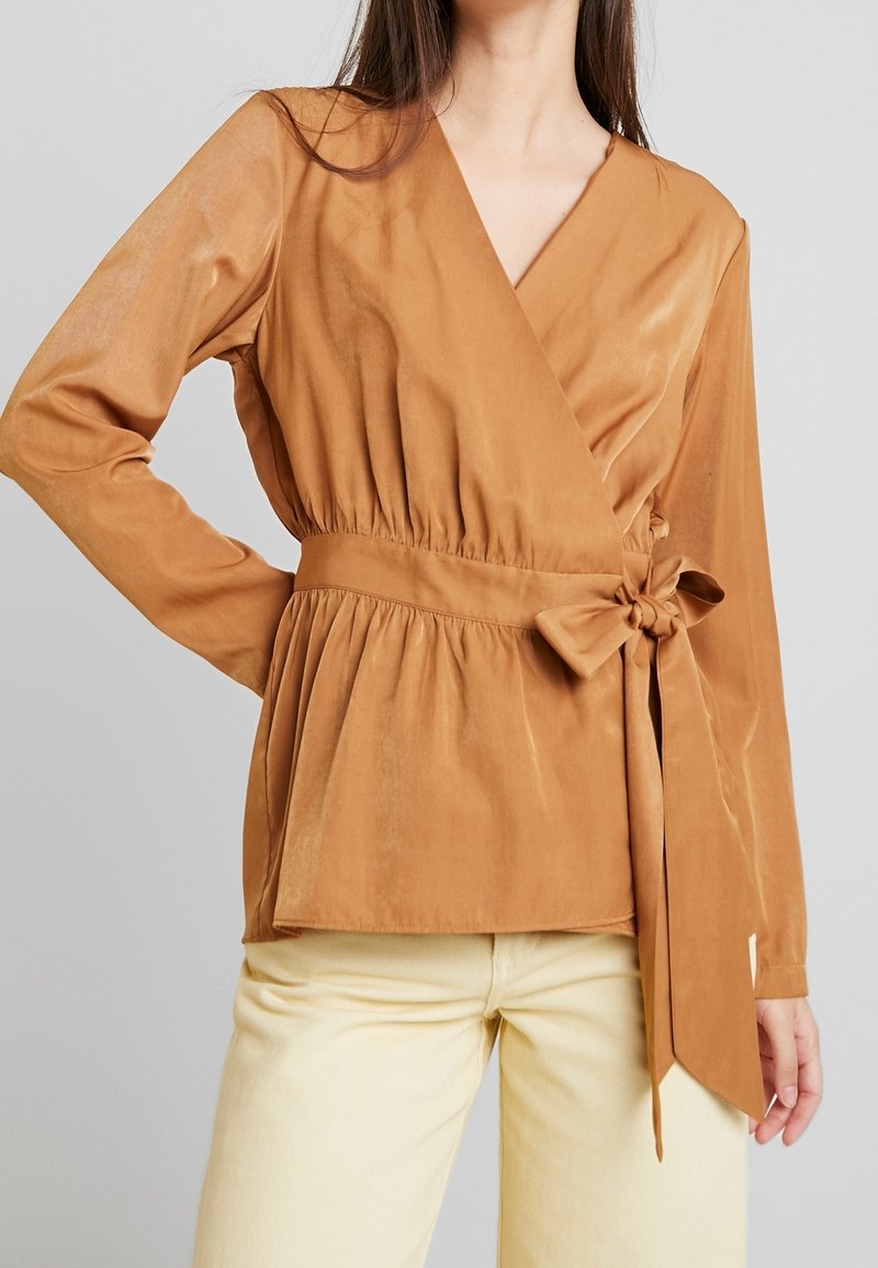 Femme portant un chemisier cache-cœur orange rouille avec un nœud sur le côté et manches longues, associé à un pantalon beige clair sur un fond gris.