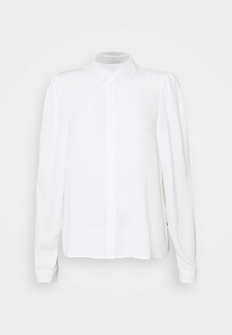 Blouse blanche à manches longues avec boutons sur le devant, col en dentelle et épaules légèrement bouffantes sur fond uni.
