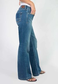 Jean en denim bleu à jambes larges avec une finition délavée, doté d'une patch en cuir marron au niveau de la taille et sur les poches avant/arrière. Texture lisse.
