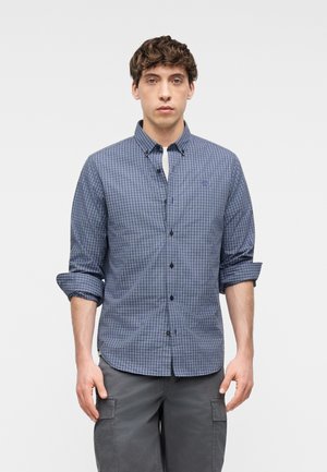 Młody mężczyzna zwrócony przodem, ubrany w niebieską koszulę w kratę z podwiniętymi rękawami oraz szare spodnie typu cargo.