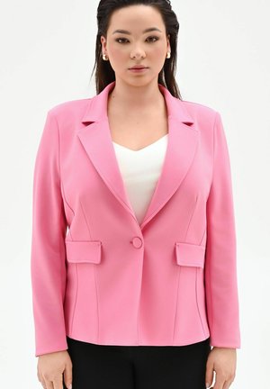 Femme portant un blazer rose ajusté sur un haut blanc et un pantalon noir, faisant face à l'avant avec une expression neutre sur un fond uni.