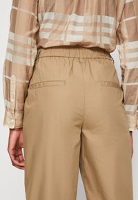 Pantalons beige à taille élastique avec poches arrière, associés à une chemise à manches longues à carreaux marron, crème et beige. Tissu en mélange de coton.