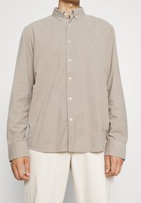 Camisa de manga larga con botones en algodón beige claro, que presenta un cuello, botones blancos y puños ligeramente sobredimensionados. Corte liso y casual.