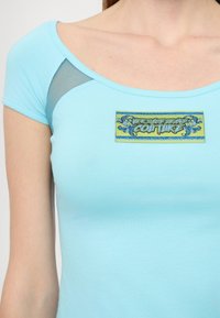Light blue short-sleeve t-shirt with a scoop neckline, featuring a decorative embroidered patch with "Versace Jeans Couture" text.