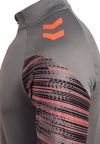 Graues Sportoberteil mit hohem Kragen, ausgestattet mit strukturiertem Stoff und einem gemusterten Ärmel in Schwarz und Rot, akzentuiert durch orangefarbene Logodetails.