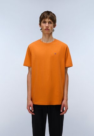 Orange Baumwoll-T-Shirt mit kurzen Ärmeln, Rundhalsausschnitt und einem kleinen Emblem nahe der Schulter. Kombiniert mit schwarzen Hosen. Schlichte, lässige Passform.