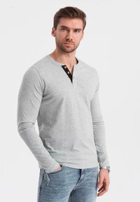 Harmaa pitkähihainen henley-paita, jossa on musta neppari ja kolme ruskeaa nappia. Pehmeää puuvillakangasta, istuva malli, yhdistetty vaaleansinisten farkkujen kanssa.