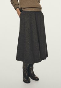 Kvindelig iført en brun sweater, nederdel midi med mørke tern og lommer, samt sorte læder støvler til midt på læggen, stående mod en ensfarvet baggrund.