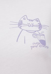White t-shirt featuring a purple cat illustration holding a drink, with the text "chase your dreams!" in playful font. Soft texture, casual design.