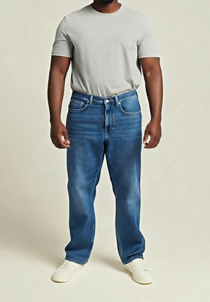 Man wearing light grey short-sleeve t-shirt, blue straight-leg jeans, and white slip-on shoes standing against plain background.