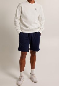 Vit sweatshirt med en liten grå logotyp på bröstet, i kombination med marinblå shorts. Modellen har på sig vita sneakers och knähöga strumpor.