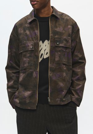 Person iført en brun og lilla camouflage lynlåstrøje over en sort sweater med beige abstrakt design og mørke bukser med tynde striber.