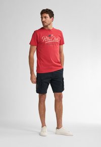 Camiseta de algodón roja con logo impreso, combinada con pantalones cortos cargo oscuros. El modelo lleva zapatillas blancas; los pantalones cortos tienen bolsillos laterales y un detalle de parche.